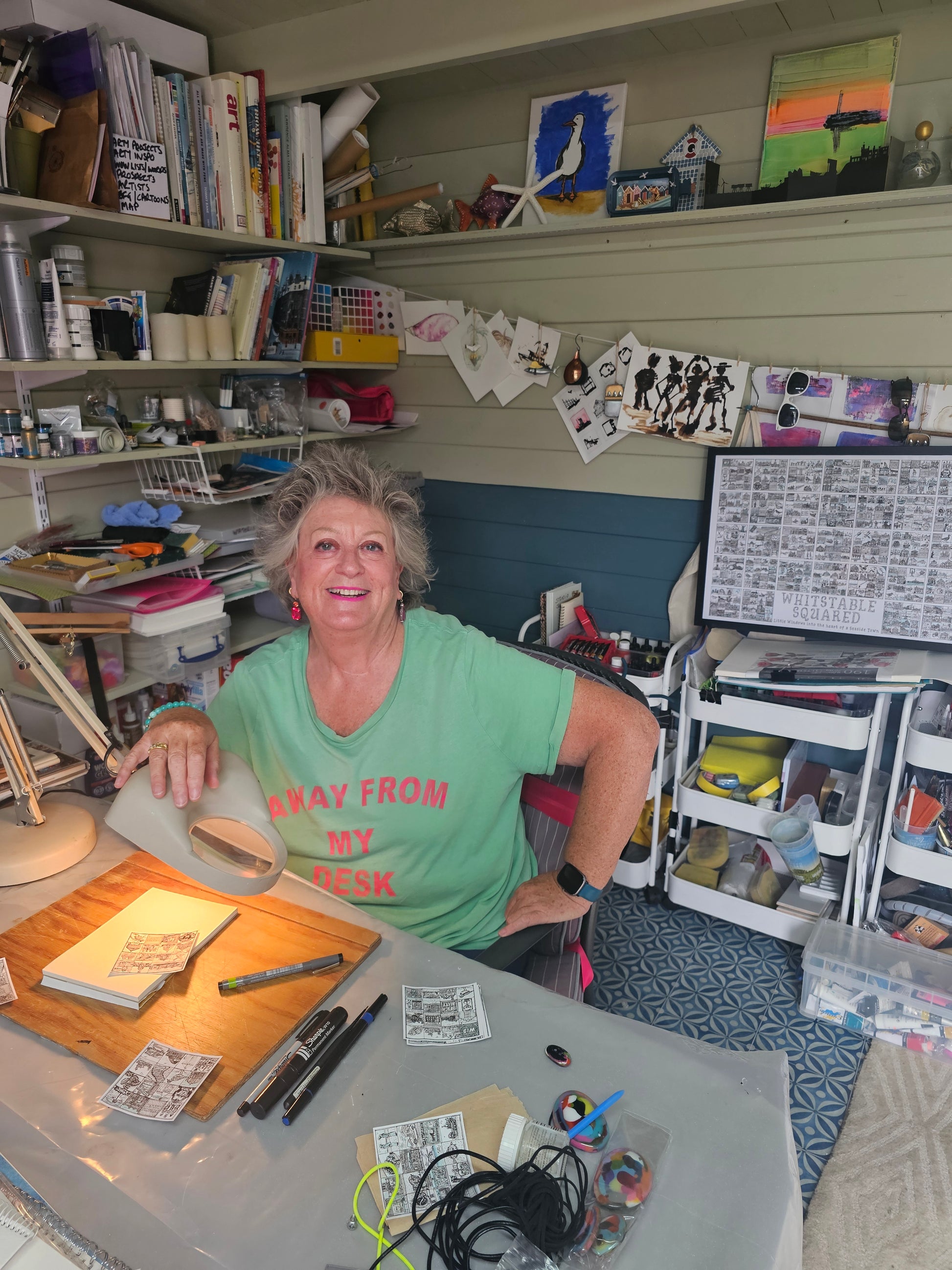 Caroline Wagstaff artist of 'Whitstable Squared' in a green shirt sitting in a her studio with books, pens and art materials around her.

' print sitting on a pink bench with her dog Boots