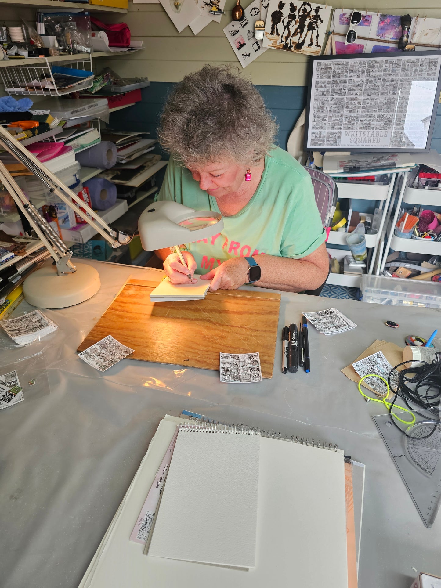 Caroline Wagstaff artist of 'Whitstable Squared' in a green shirt working at her desk sitting in a her studio with books, pens and paper around her.