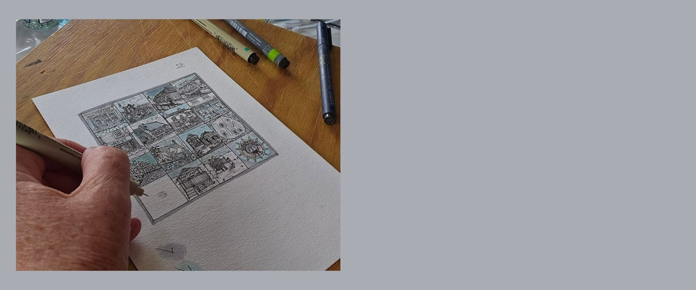 The artists hand holding a pen over a piece of paper with a black and white illustration that forms part of Whitstable Squared on a wooden table.