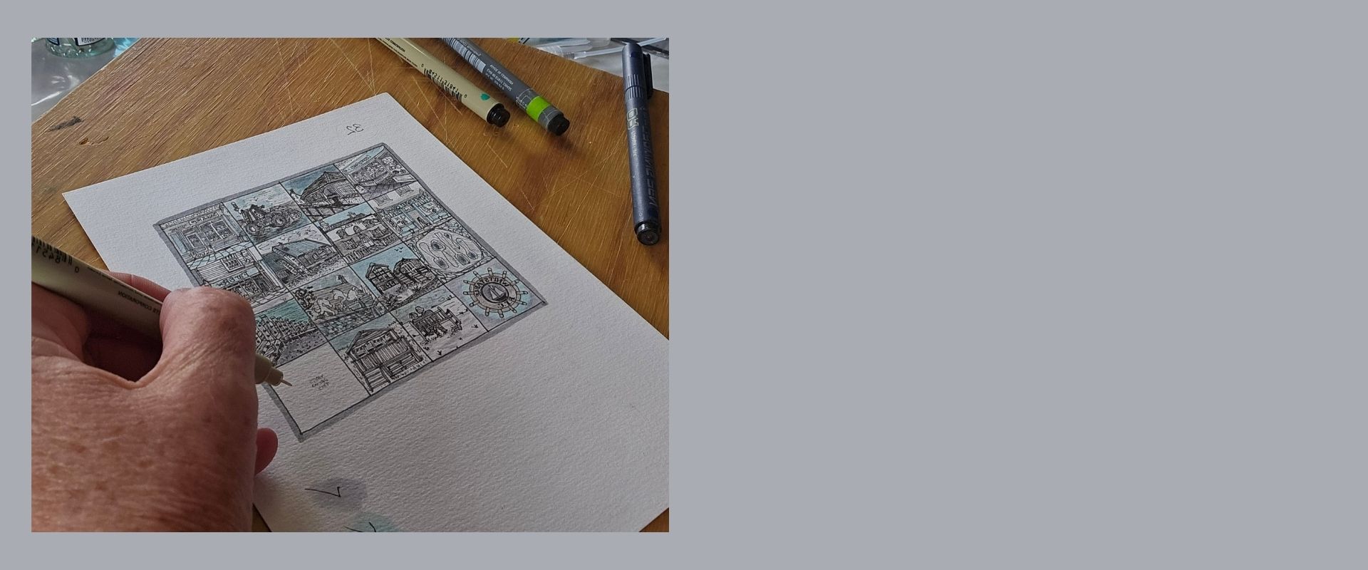 The artists hand holding a pen over a piece of paper with a black and white illustration that forms part of Whitstable Squared on a wooden table.