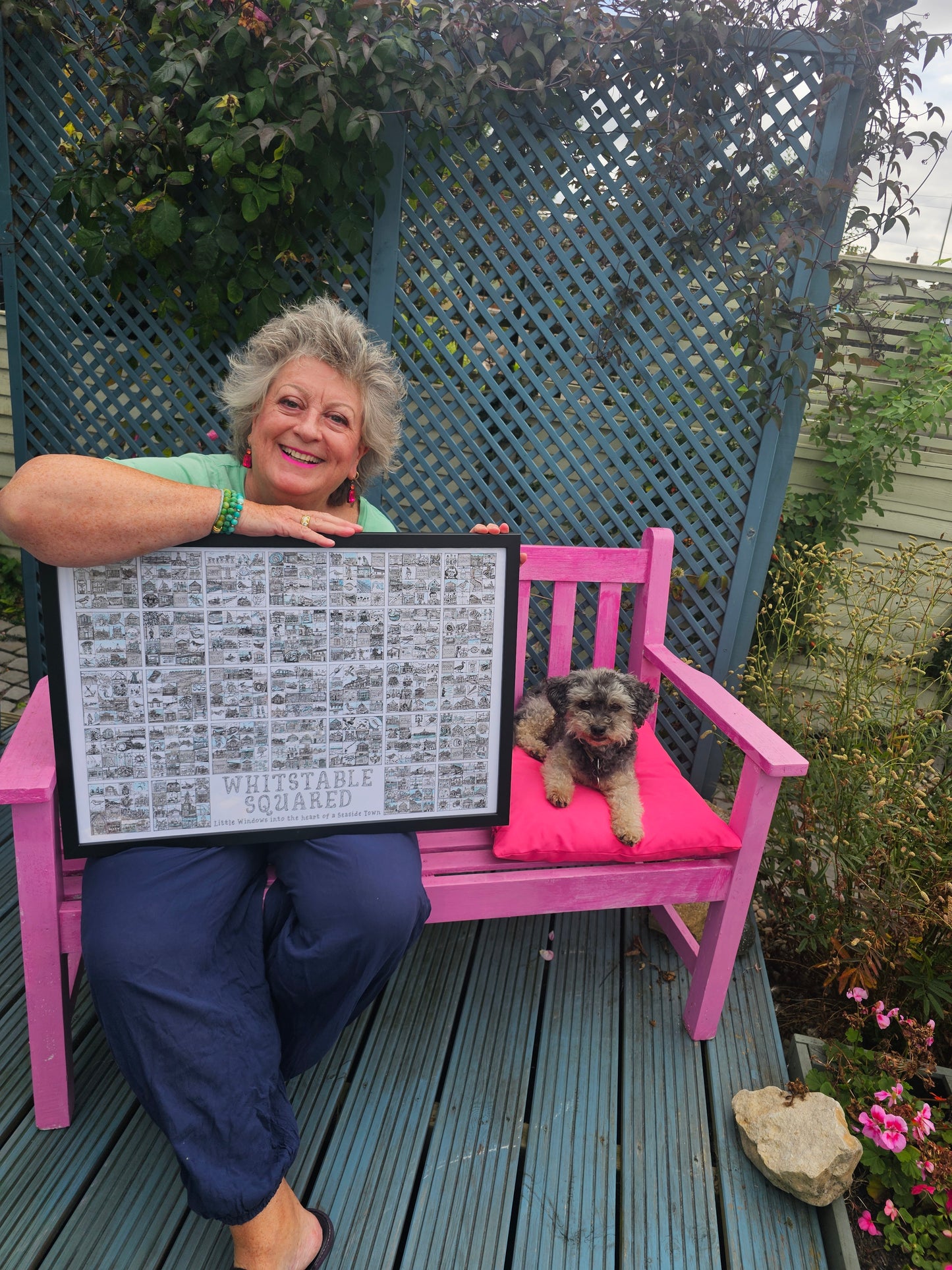 Caroline Wagstaff holding a framed print of a landscape format 'Whitstable Squared' print sitting on a pink bench with her dog Boots